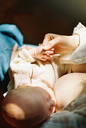 A newborn baby in a knit onesie gently holds an adult's finger in soft warm sunlight.