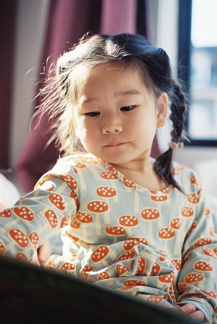 Young Asian girl in mushroom-print pajamas reading a book in warm sunlight.