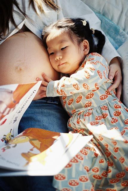 Young Asian toddler girl in mushroom print dress resting head on pregnant mother's belly while reading a book together.