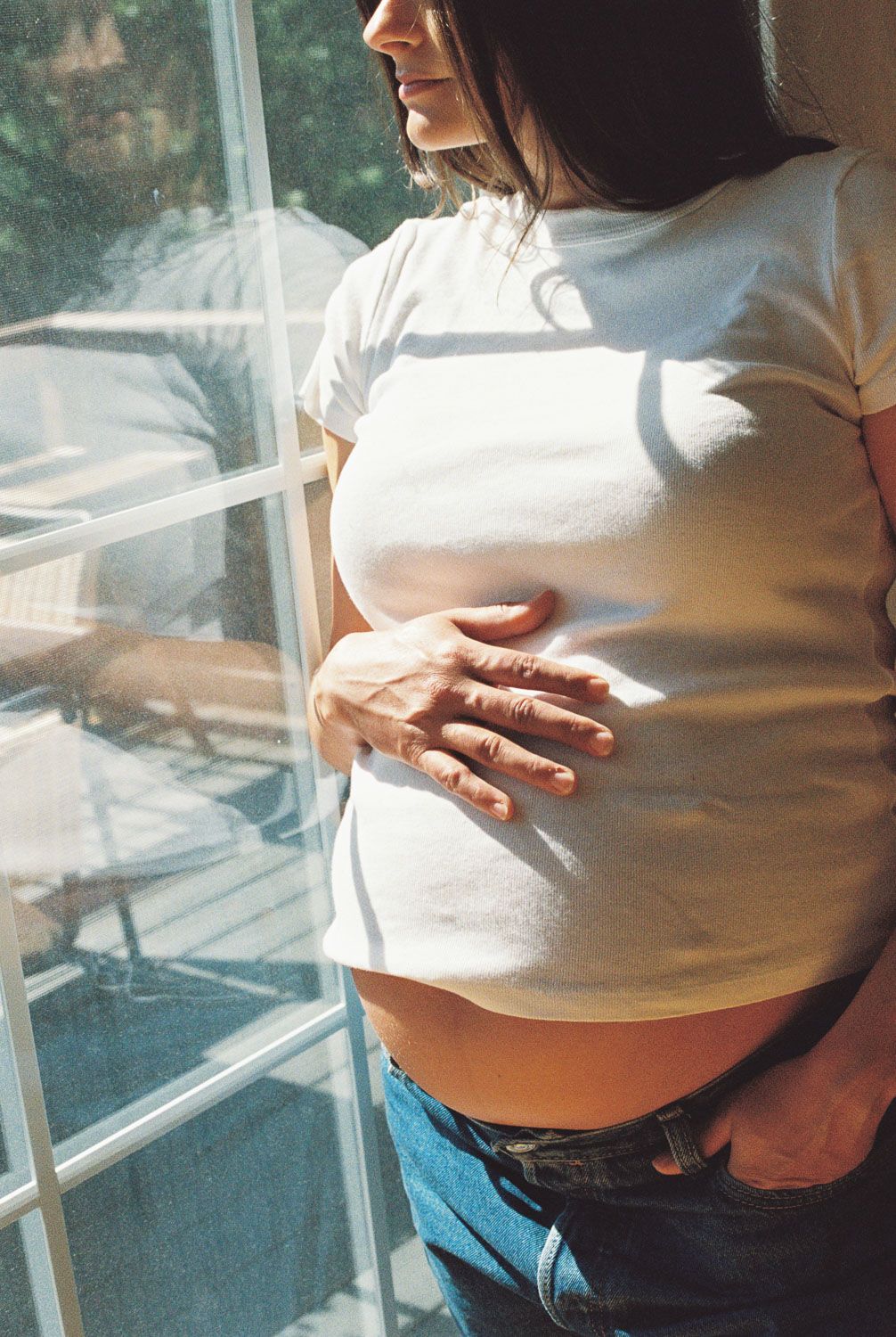 Close-up of pregnant woman in white tee cradling baby bump near sunny window in jeans.