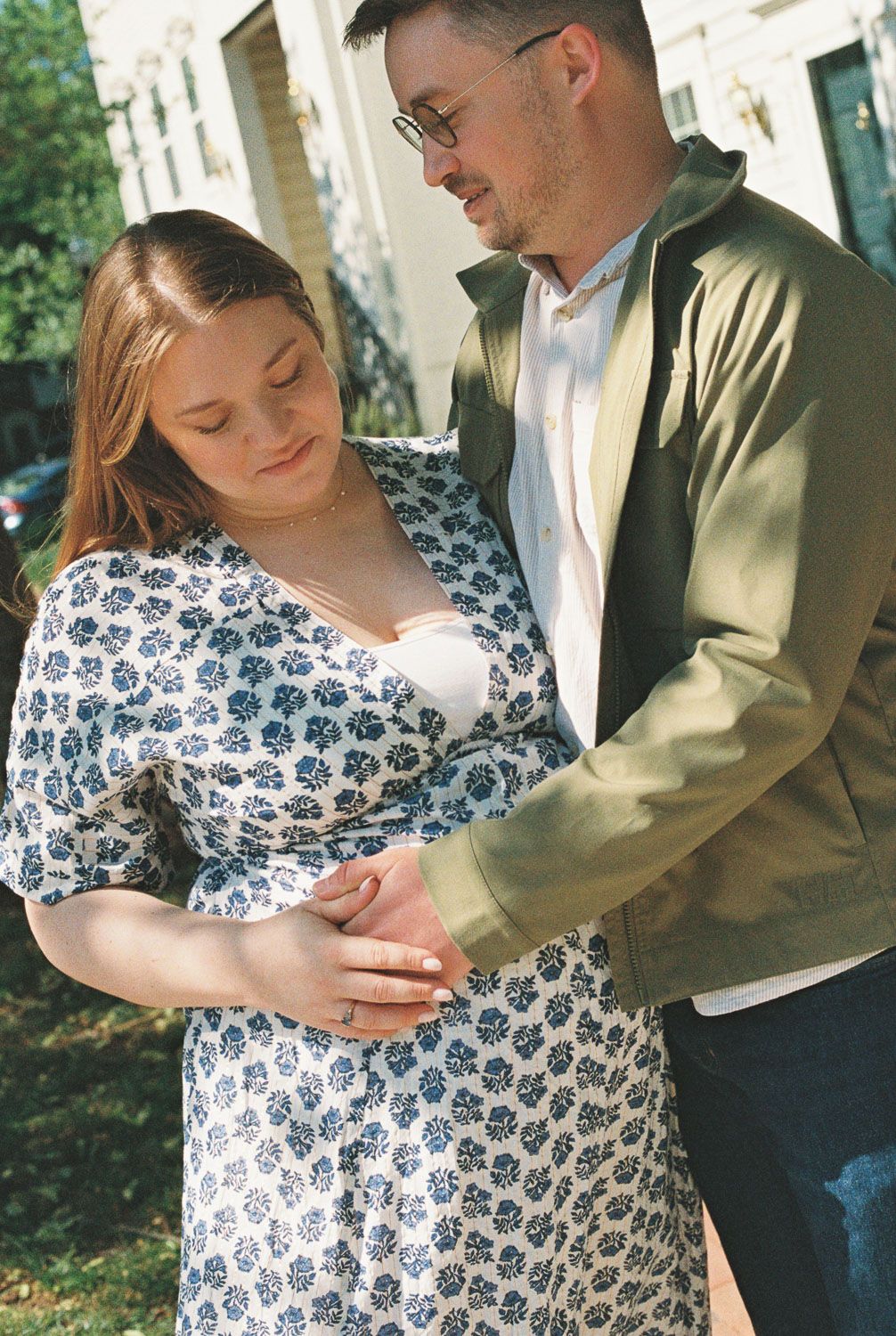 Couple tenderly cradles pregnant belly outdoors, woman in floral dress, man in olive jacket, sharing an intimate moment.