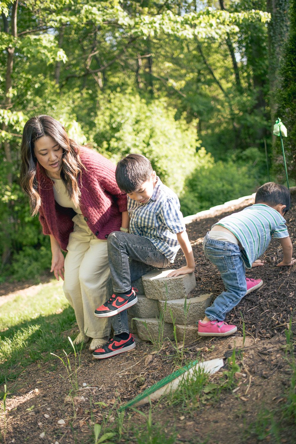 A mother helps her two young children play on stone garden steps surrounded by lush green trees outdoors.