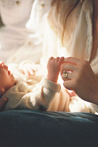 A mother gently holds her newborn baby's tiny hand, wearing a ring, in a soft warm light.