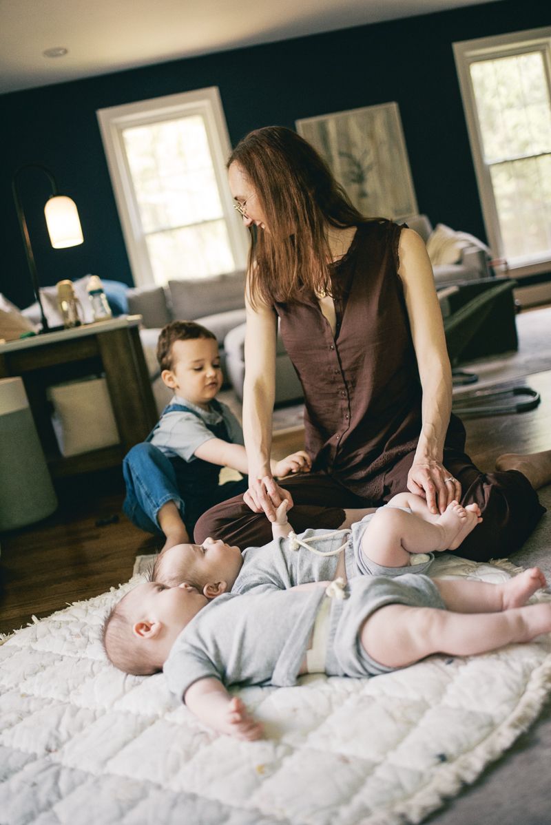 A mother sits on a white rug playing with her two young babies and toddler son in a cozy living room.