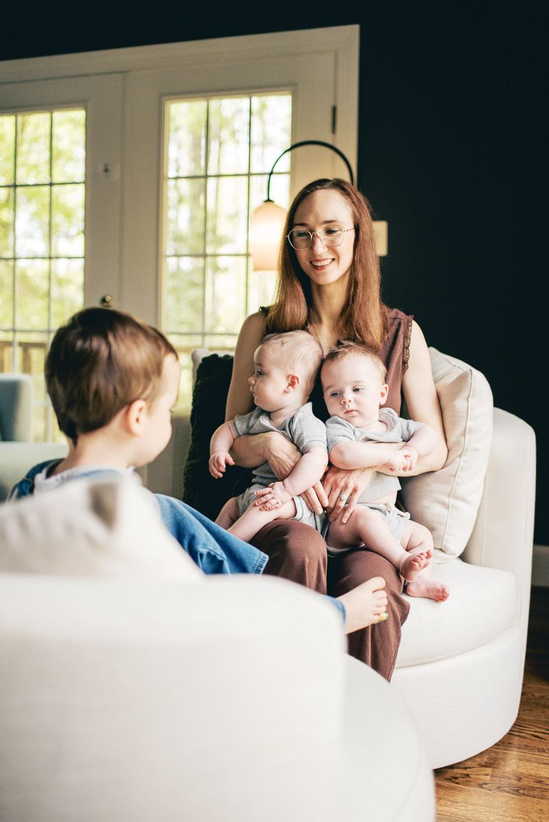 A smiling mother sits in a white chair holding twin babies while an older child sits nearby in a cozy living room.