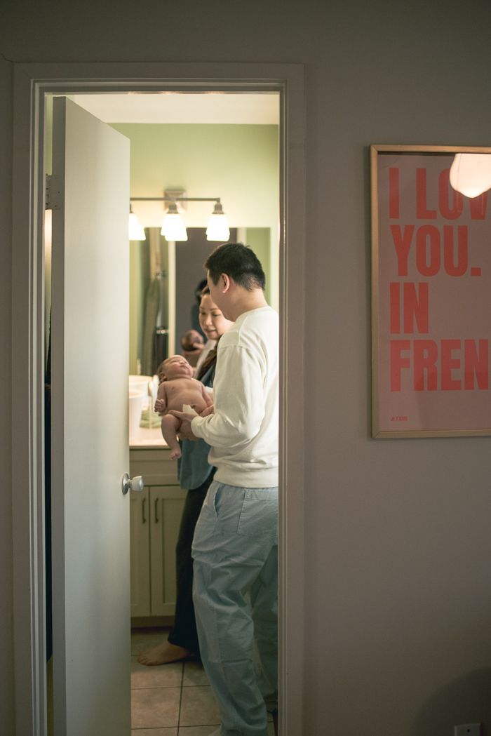 Parents tenderly holding their newborn baby in a cozy bathroom doorway, with a decorative wall print nearby.