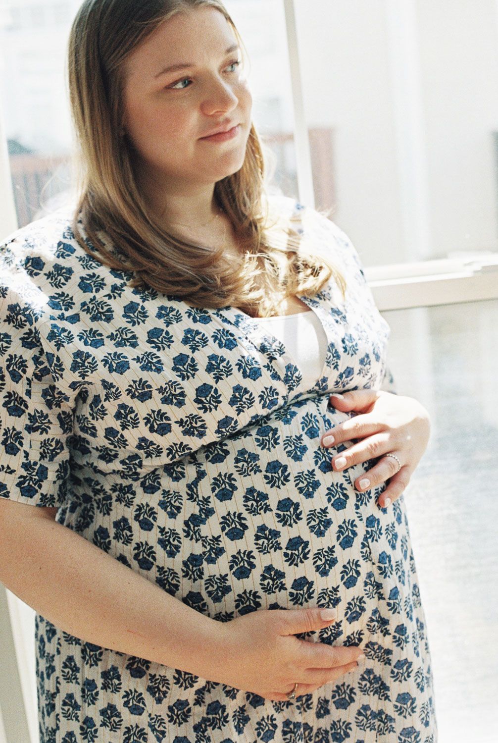 Pregnant woman in blue floral dress cradling baby bump, standing near window with soft natural light.