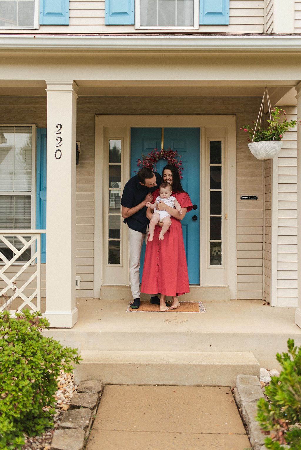 Family stands together on the front porch of a beige house with blue shutters and decorative red wreath.