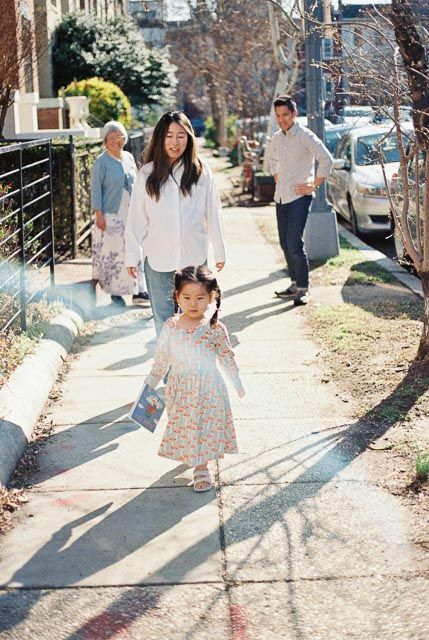 Asian family walking on a sunny sidewalk, with a young girl in a floral dress leading ahead of her parents.