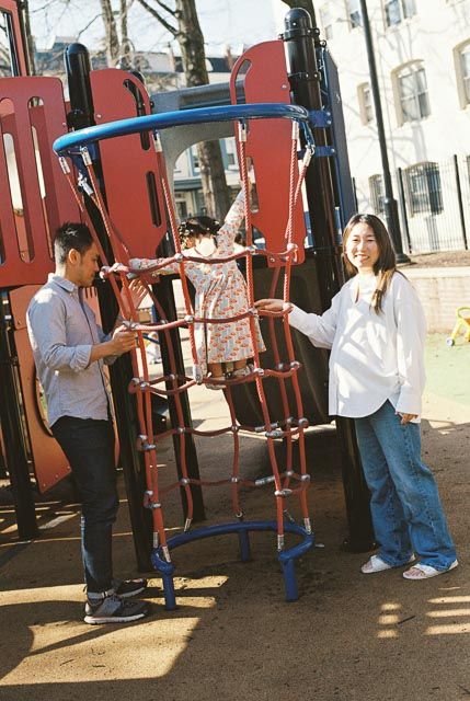 Two adults assist a child climbing a red and blue playground structure on a sunny day.