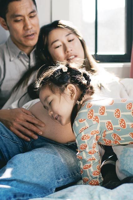 Asian family resting together on bed, young girl sleeping on mother's pregnant belly while father watches lovingly.