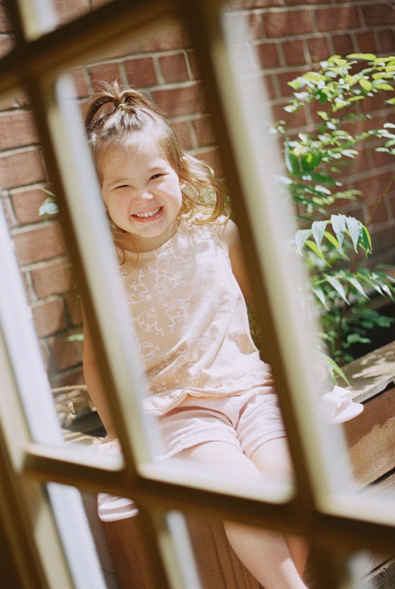 Smiling young girl with a messy updo sits outdoors in a light pink outfit, framed by a wooden window with brick wall and green leaves.