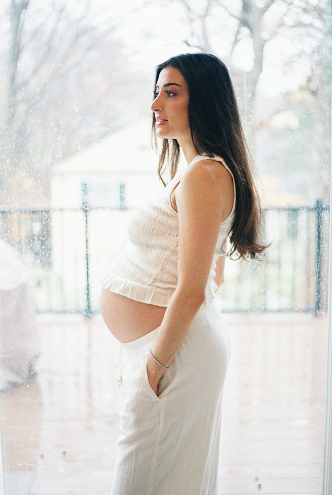 A pregnant woman in a white dress poses in profile by a bright window during a maternity photoshoot.