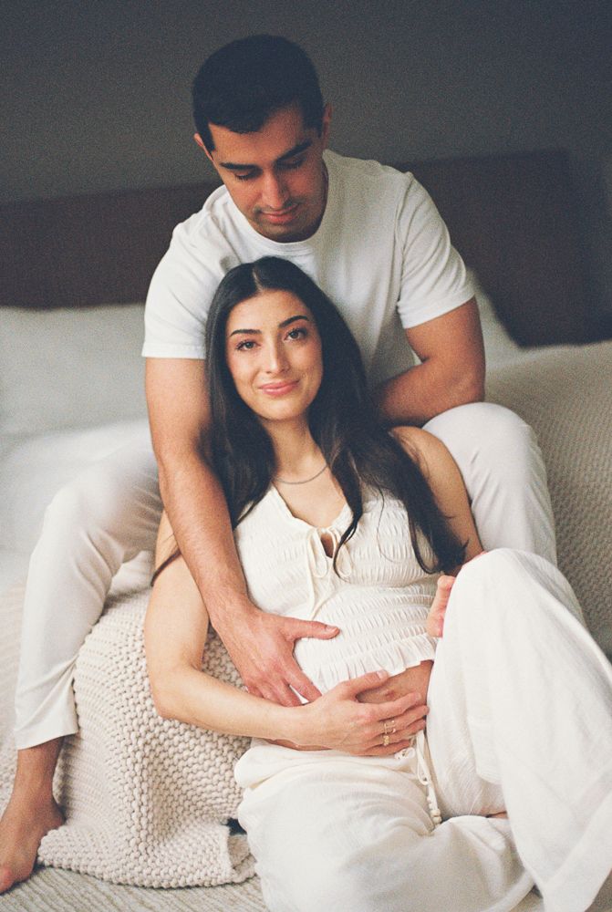 A couple in white clothing sits together intimately on a couch during a maternity photoshoot.