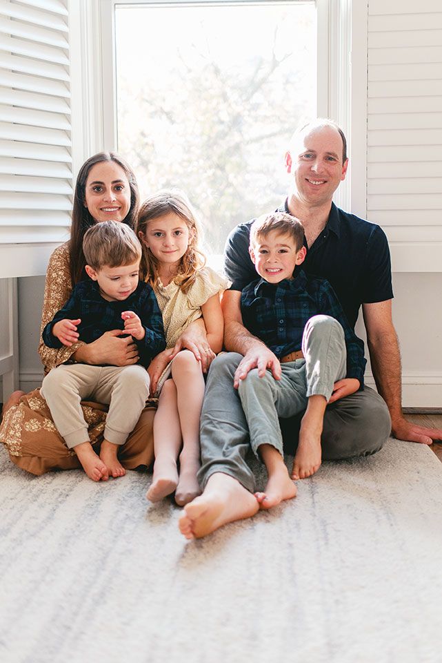 Family sitting together on white carpet in bright sunlit room with plantation shutters in background.