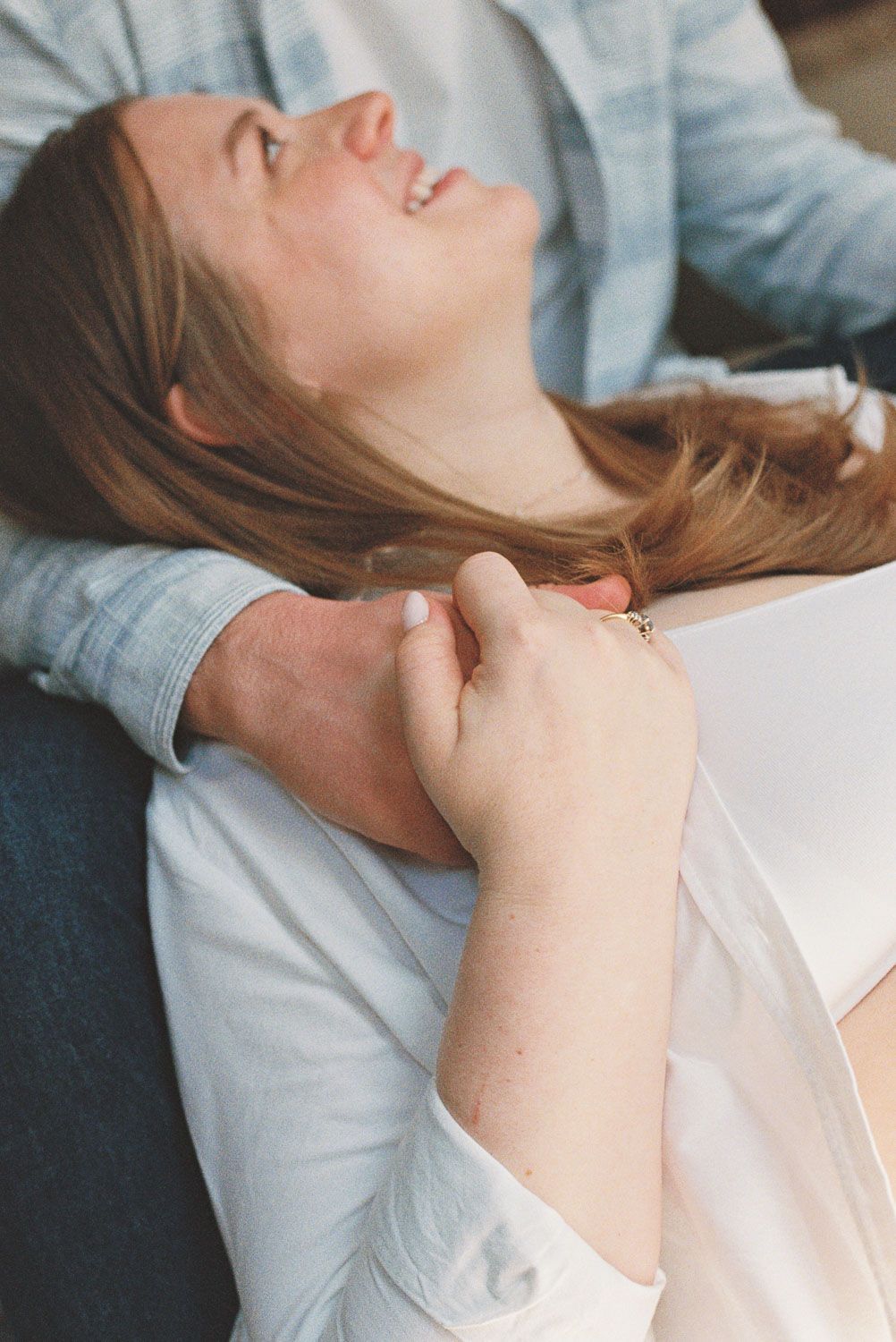 A laughing woman with long hair leans back joyfully while a partner tenderly holds her shoulder during a maternity session.