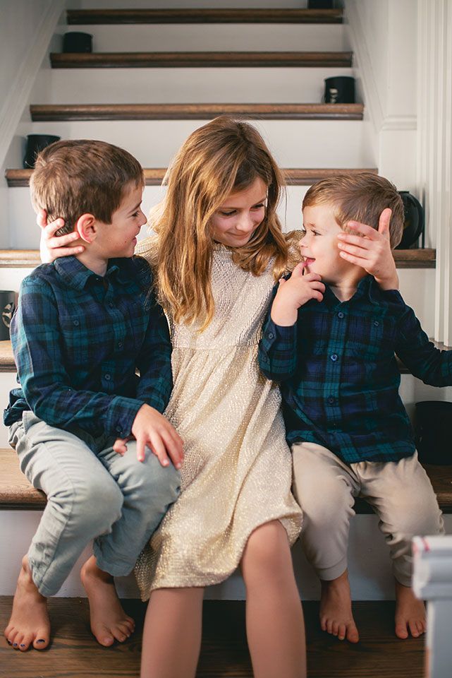 Three siblings share a playful moment together on wooden stairs, wearing coordinated outfits of plaid and cream.