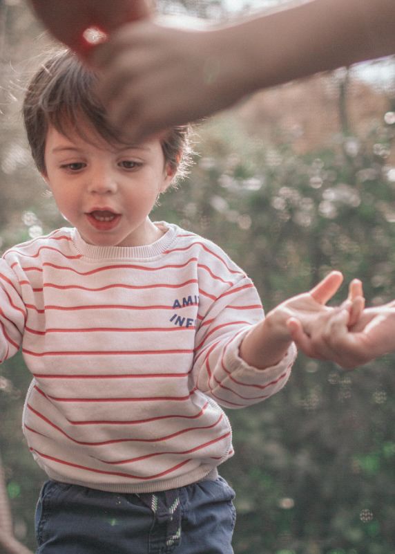 A toddler wearing a red and white striped shirt plays joyfully in a garden setting.