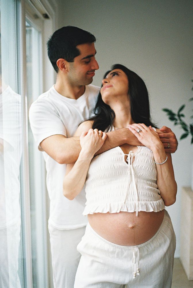 A couple in white clothing embraces tenderly in a minimalist room with natural light streaming through windows.