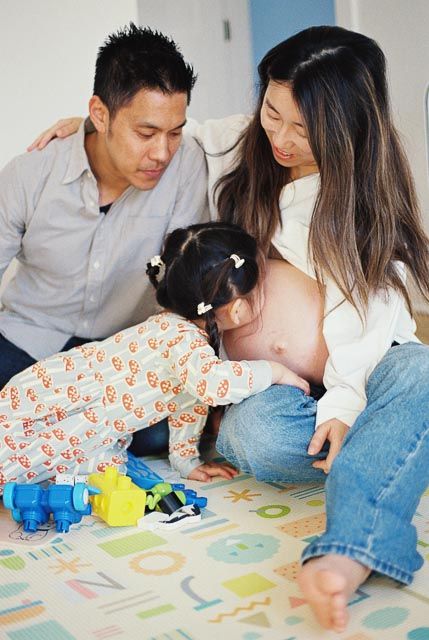 Pregnant Asian woman and man watch their young daughter play with toy robots on a colorful floor mat.