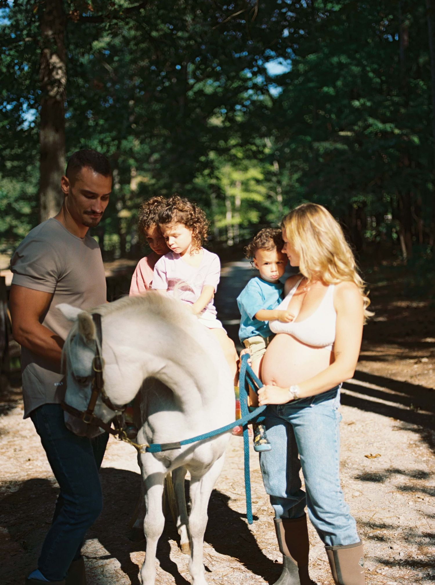A family spends time with a white pony on a wooded trail during a sunny day.