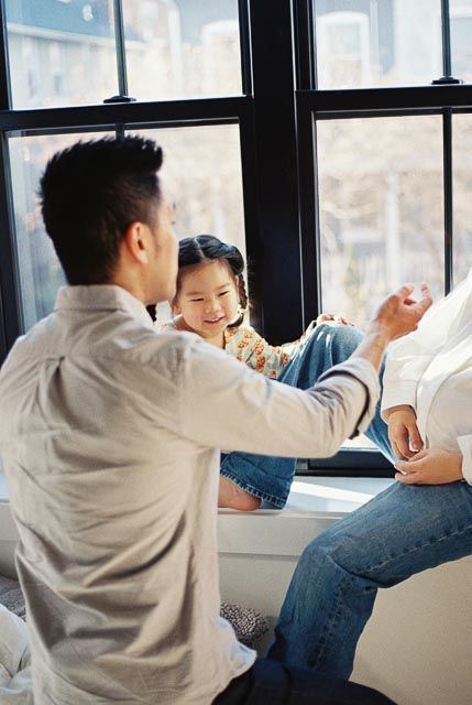 Asian father playfully interacts with smiling young daughter sitting on windowsill in bright modern home.