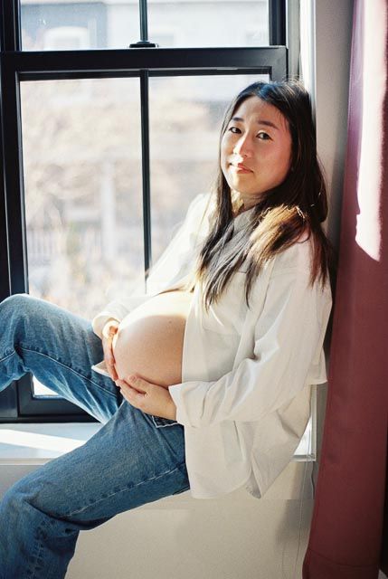 Pregnant woman in white blouse and jeans sitting by a sunlit window, cradling her baby bump.