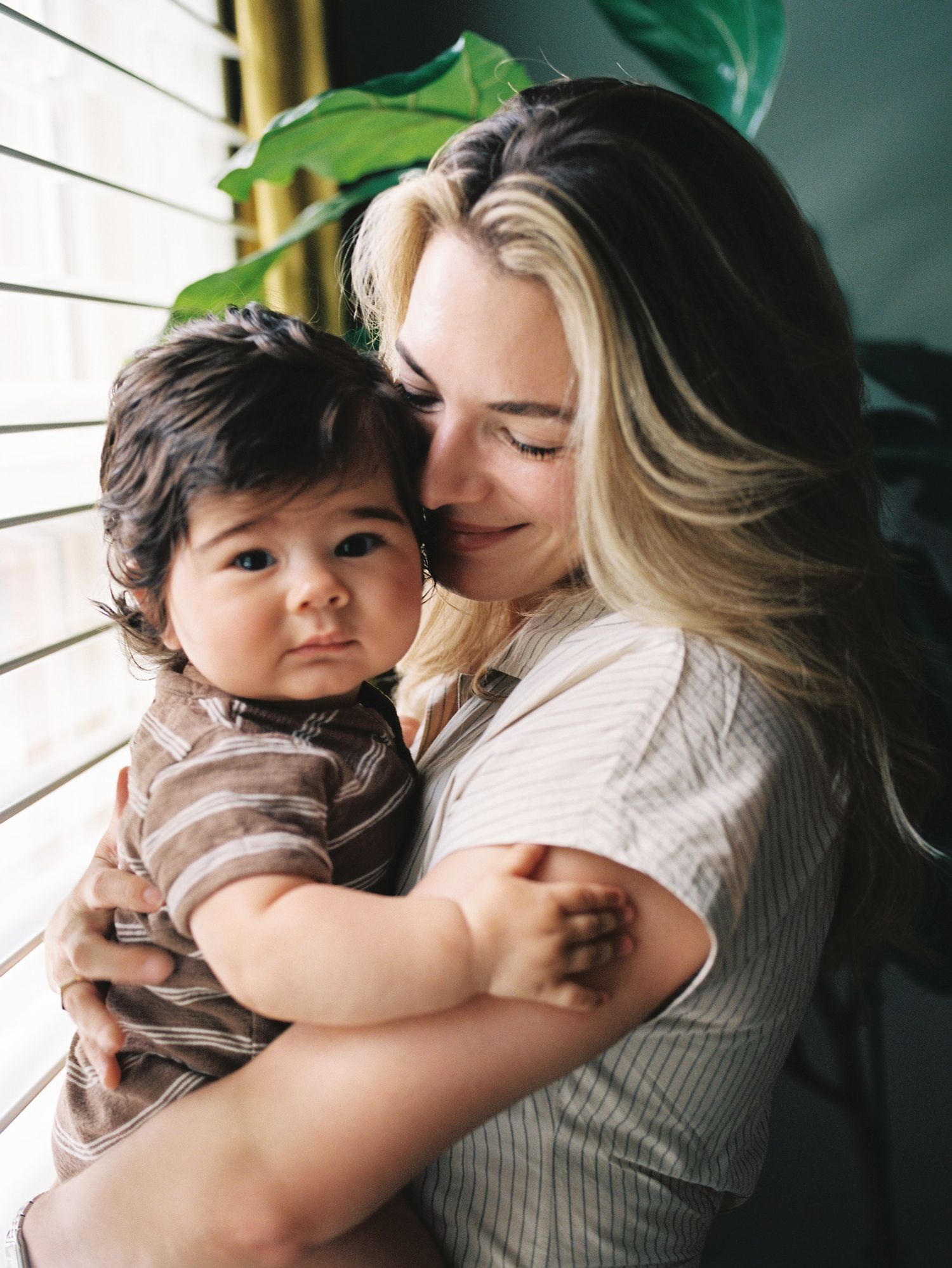Tender embrace captured showing an intimate parental bonding moment by window with green plant in background.