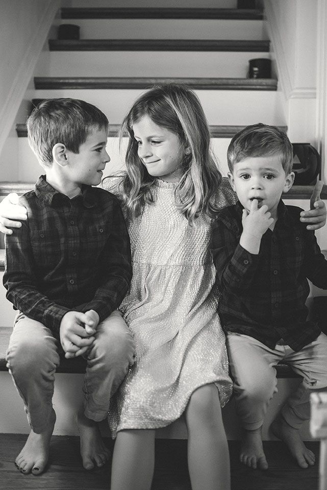 Three siblings sit on wooden stairs sharing a candid black and white moment together.