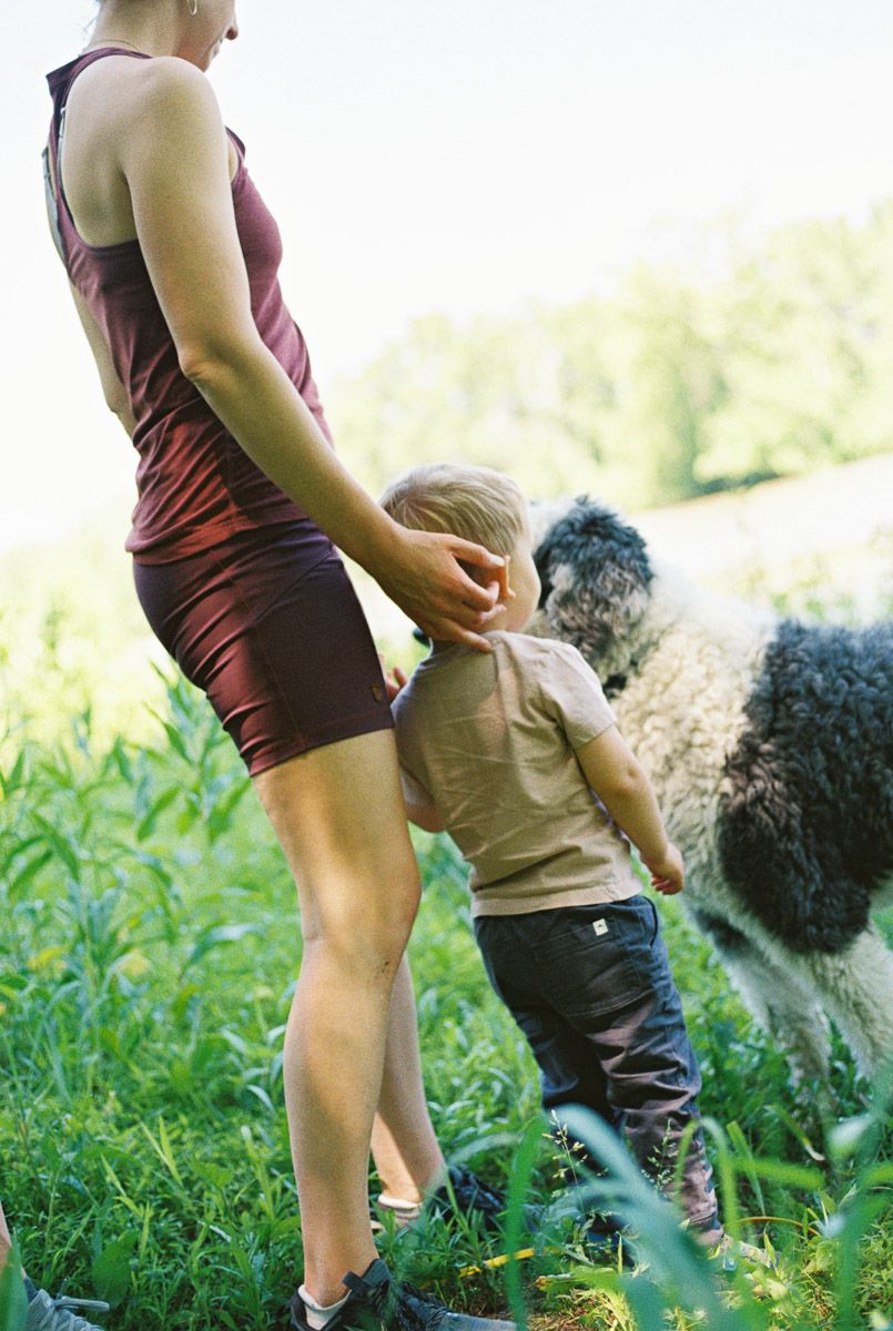 A woman in a red dress holds hands with a toddler beside a fluffy dog in a lush green field.