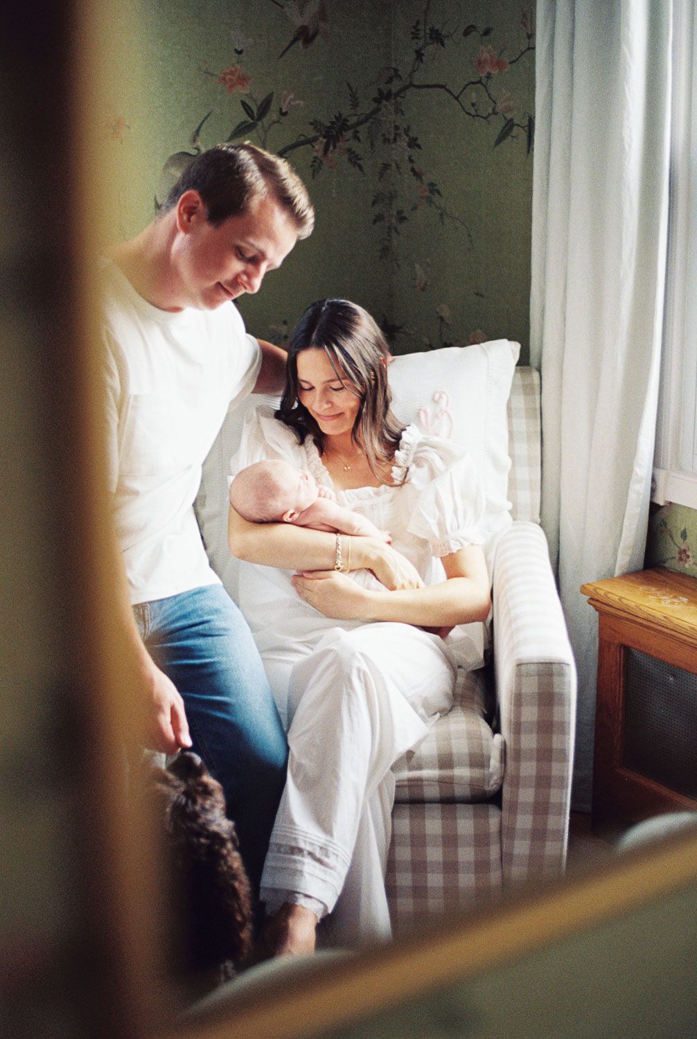 New parents admire their newborn baby in a cozy nursery, mother seated in a plaid armchair with a small dog nearby.
