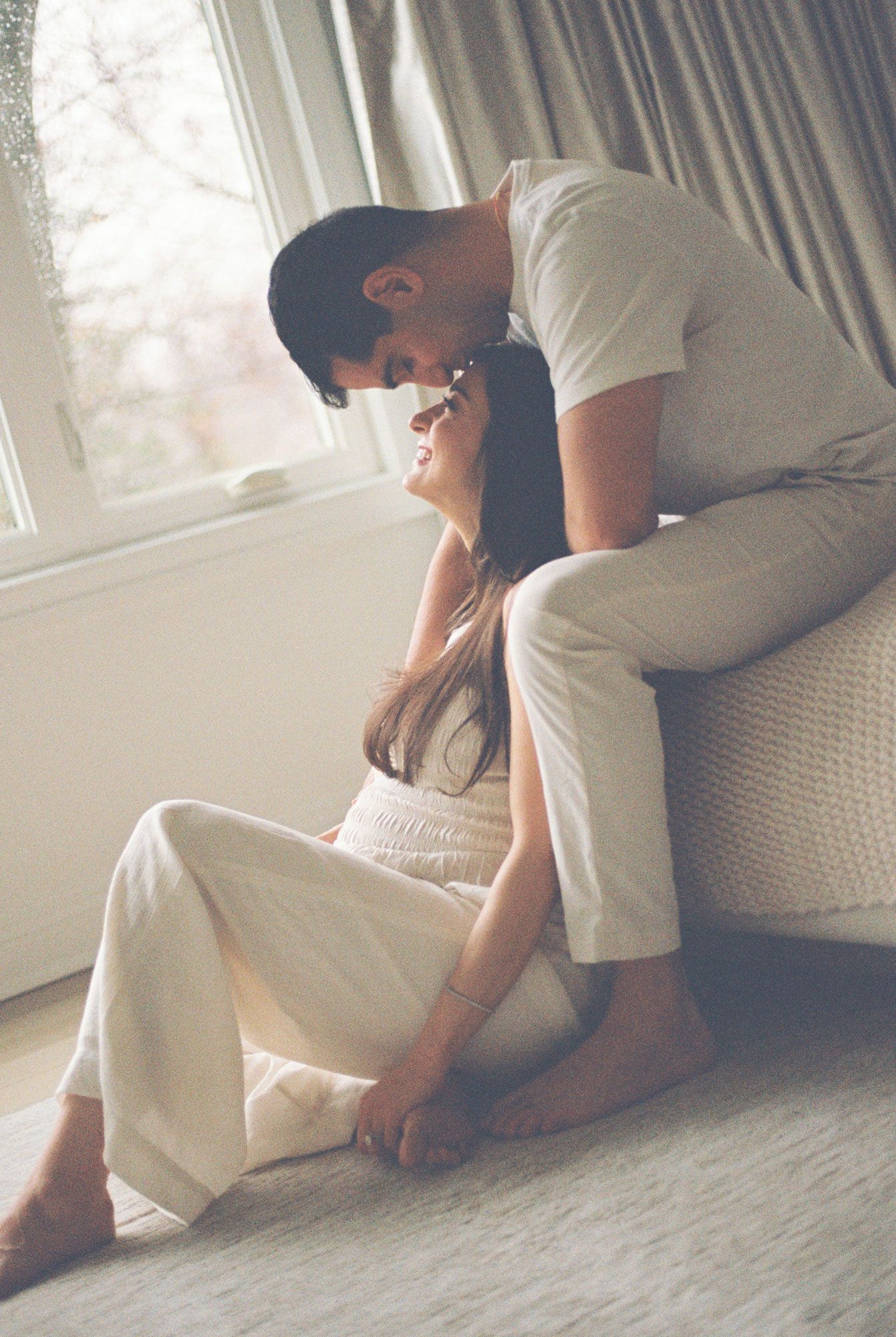 Couple in cream outfits share an intimate moment by a bright window, the man leaning over the woman as she sits on a soft rug.