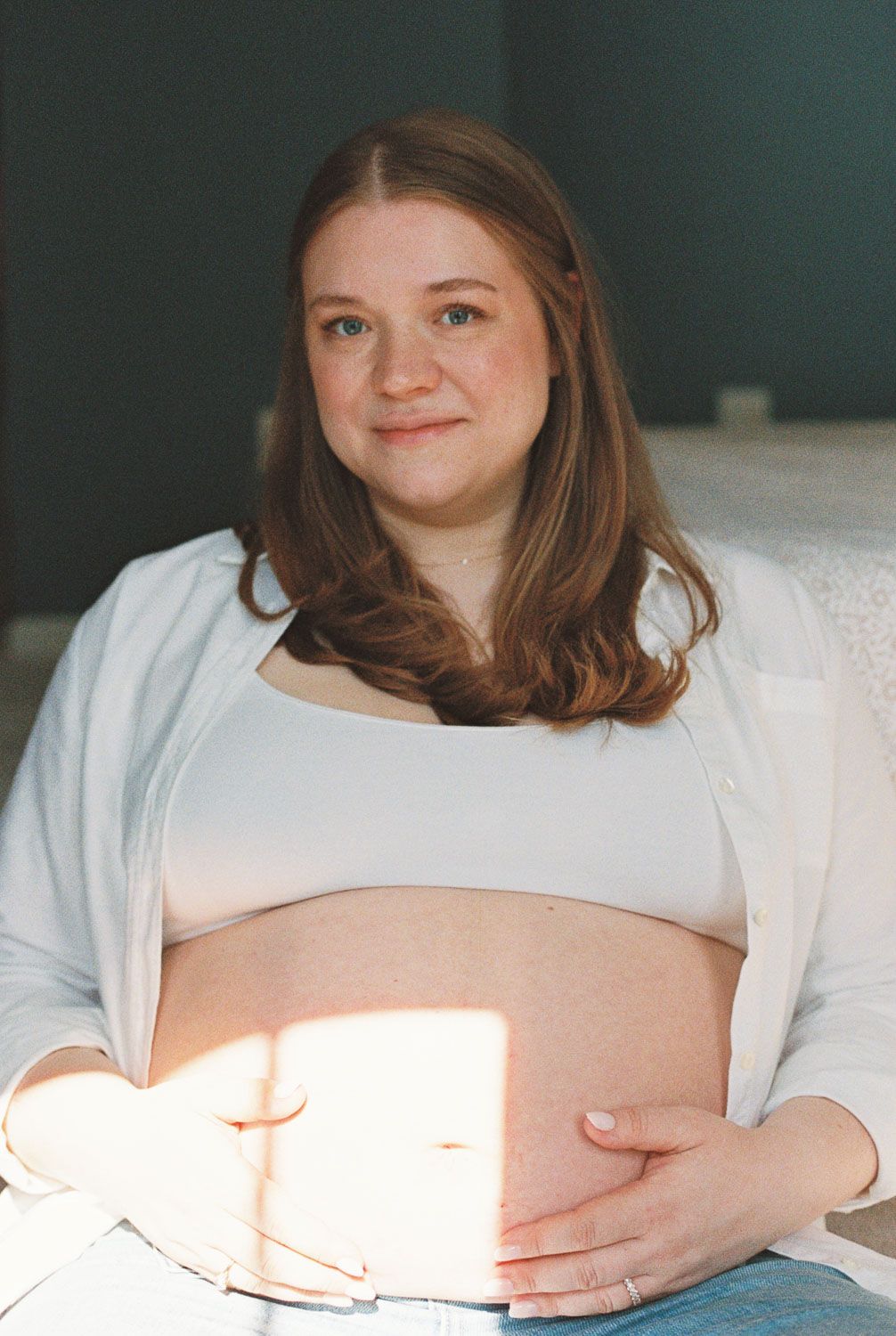 Pregnant woman in white crop top and cardigan cradling her baby bump while sitting in soft natural light.