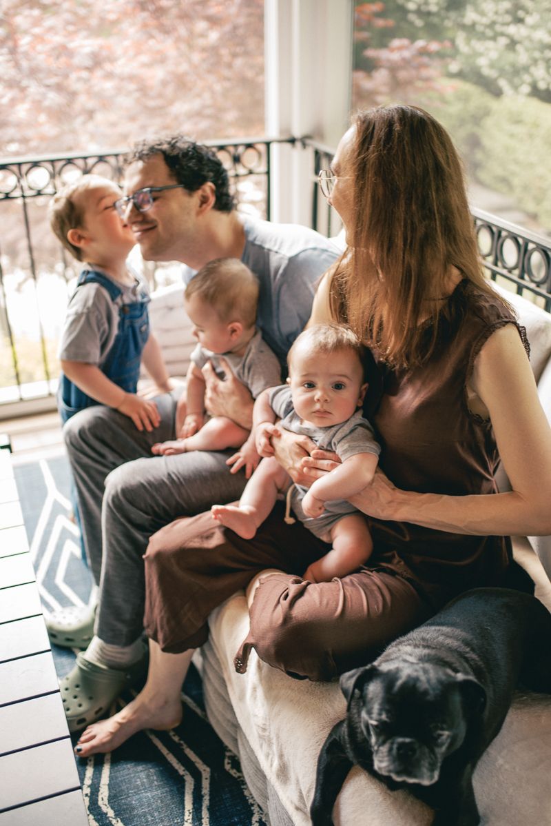 Family with two babies and toddler sitting on porch with black dog, enjoying a warm indoor moment together.
