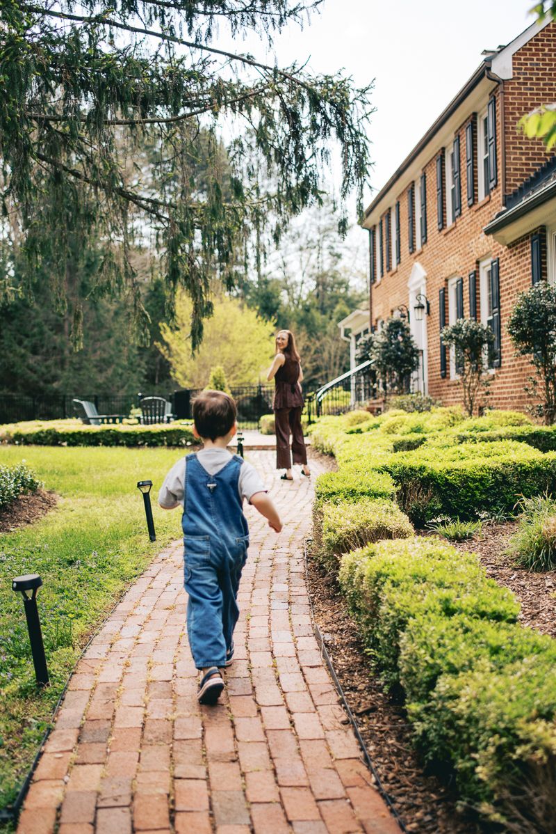 Young boy in denim overalls running toward mother on brick garden path.