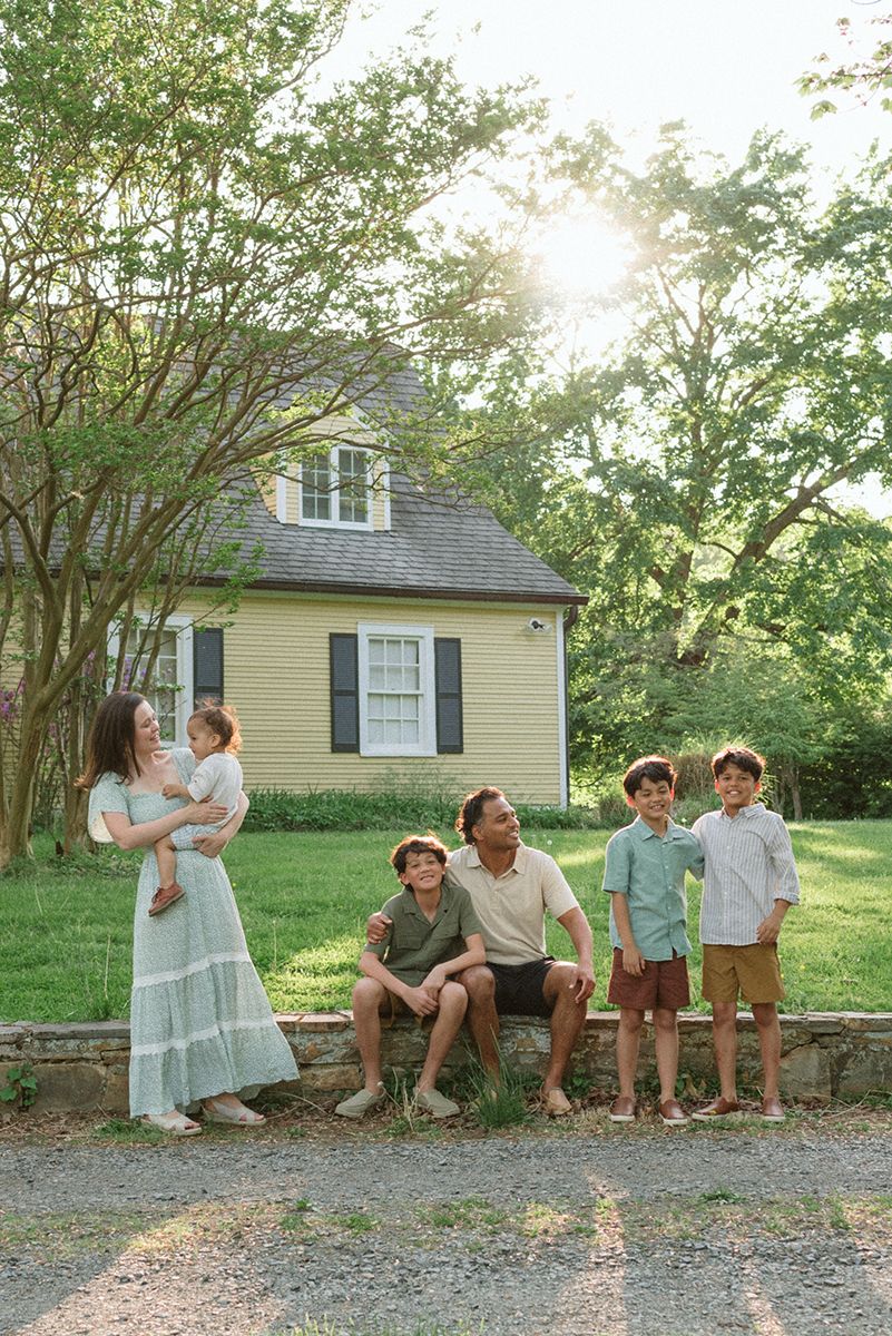 A group gathers in front of a yellow cottage house with grey shutters at sunset.