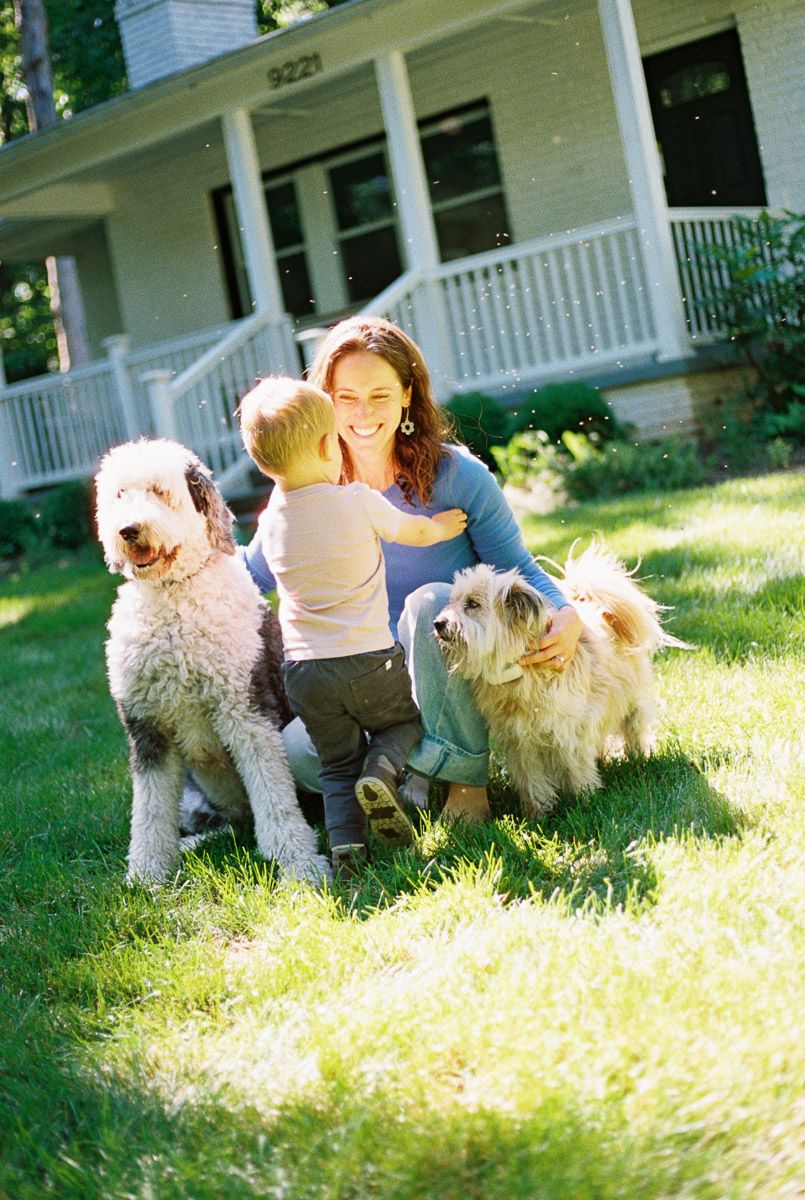 Woman smiling with toddler and two fluffy dogs on green lawn in front of white house with porch.