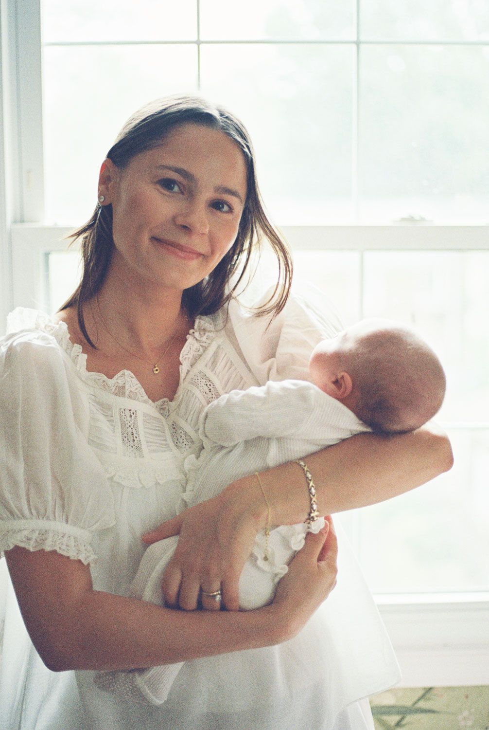 Smiling brunette mother in white lace dress holds sleeping newborn baby near a sunlit window.