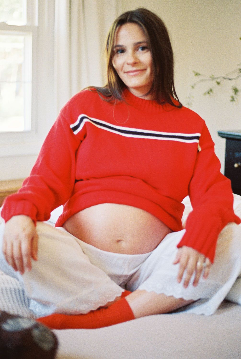 Pregnant woman in red striped sweater sitting cross-legged on bed, showing baby bump with a gentle smile.