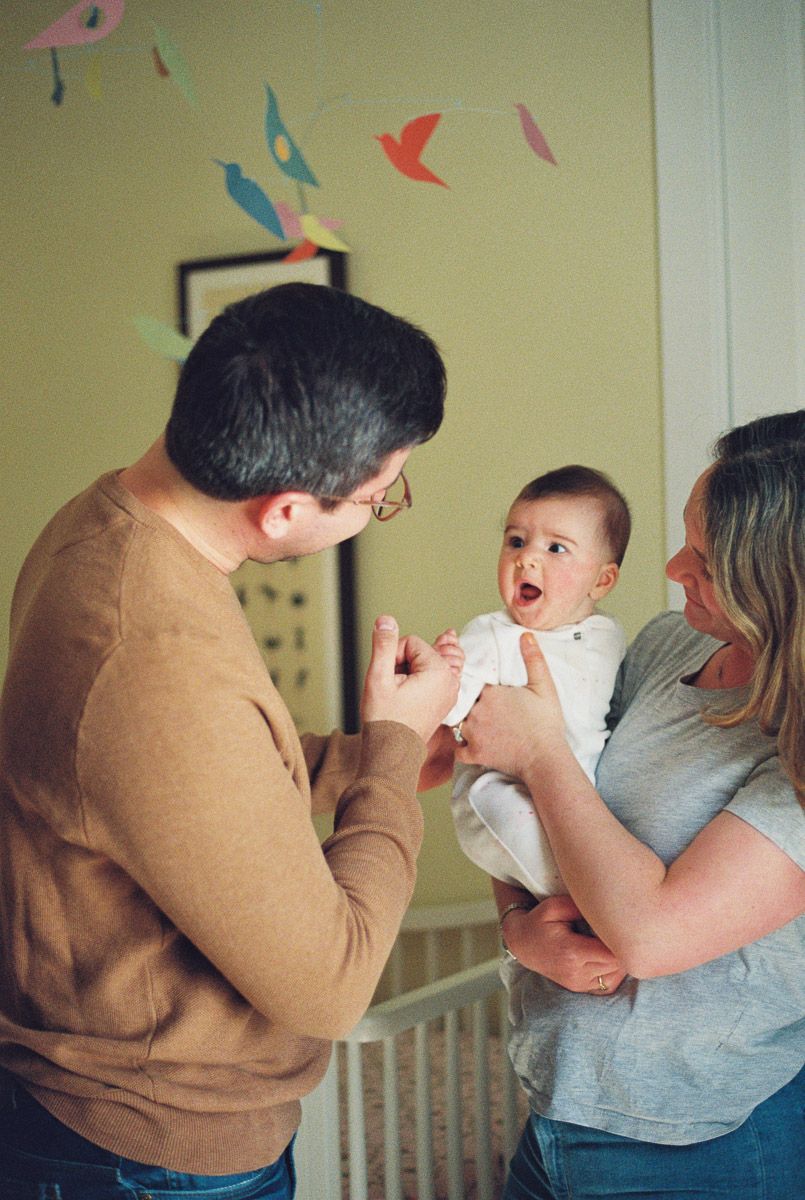 A smiling father and mother play with their wide-eyed baby beside a crib in a nursery with a colorful paper bird mobile overhead.