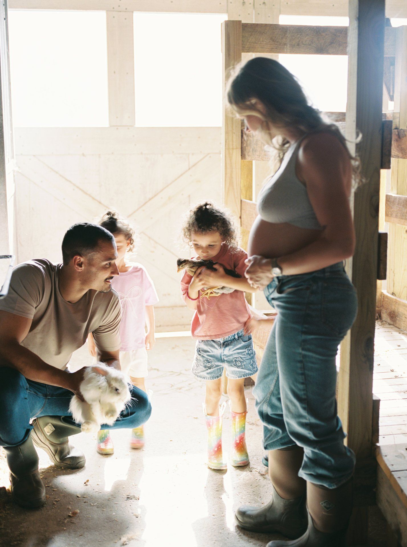 A family gathers in a rustic barn setting with a small animal, creating a warm moment together.