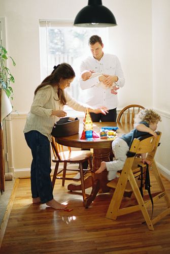 Young family with a newborn and toddler in a high chair gathered around a wooden dining table indoors.