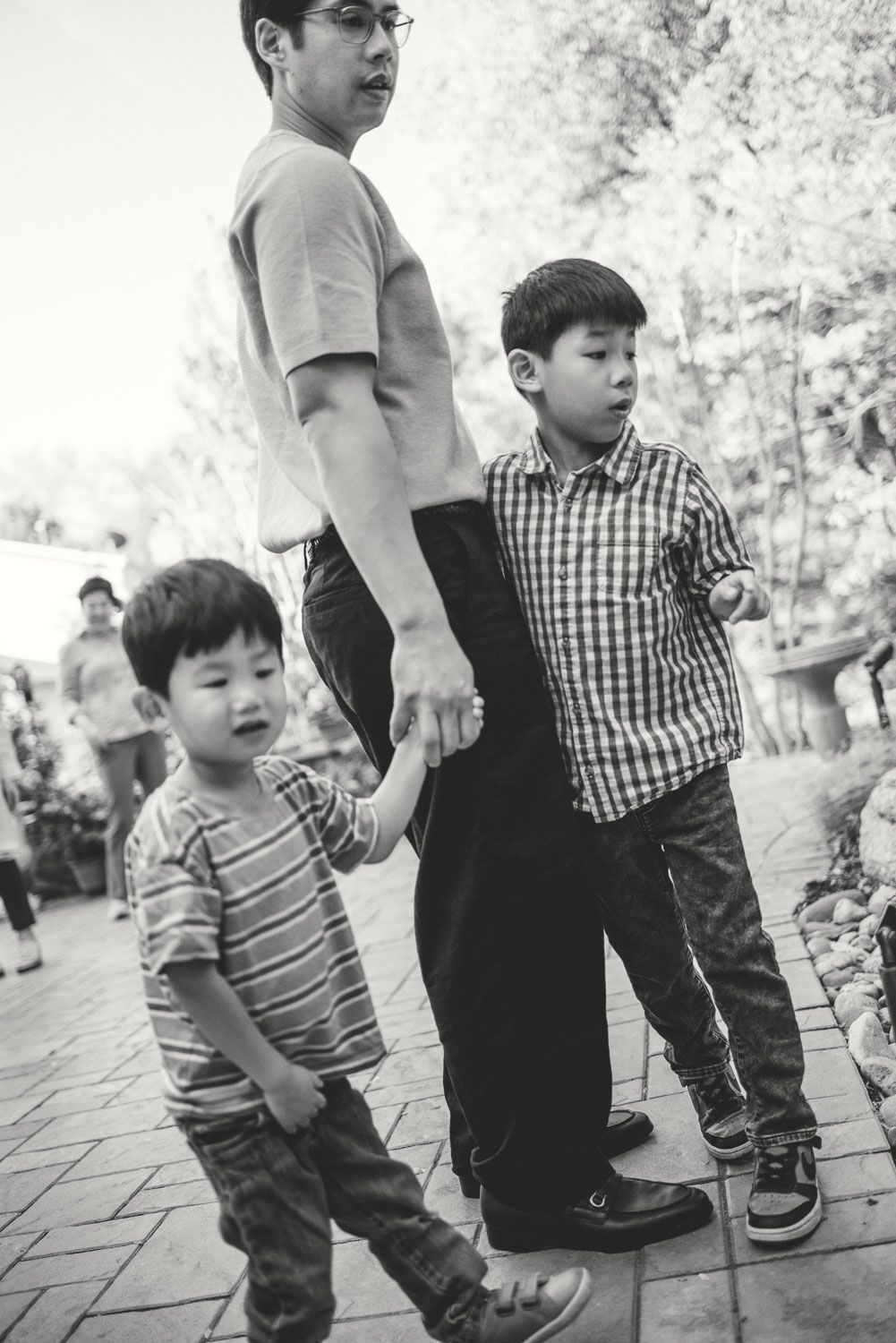 Black and white photo of a father walking with two young boys holding hands on a brick pathway outdoors.