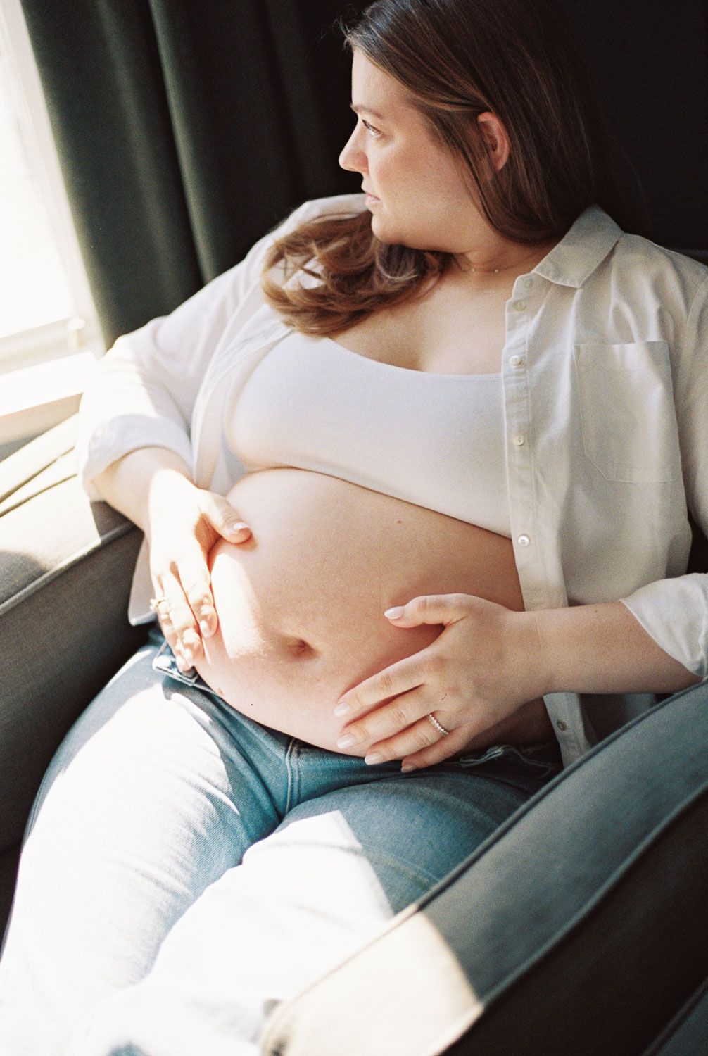A pregnant woman sits in a chair by a sunlit window, cradling her bare baby bump with both hands.