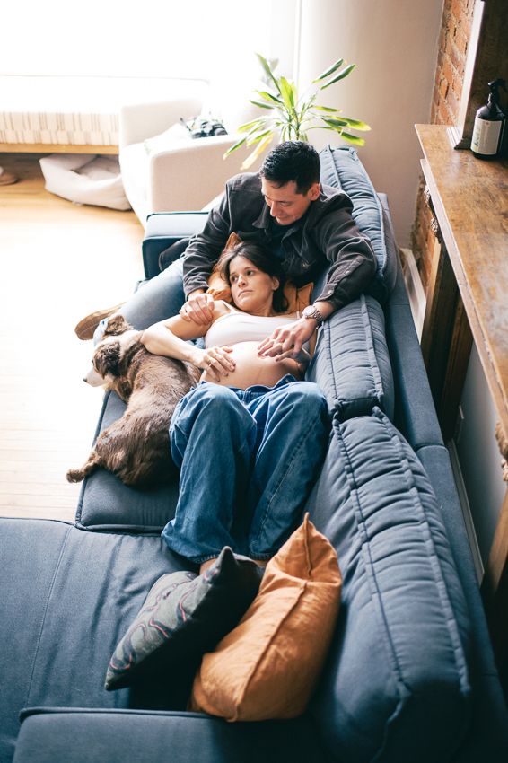 A couple relaxes together on a gray couch with their dog in a cozy living room with natural light.