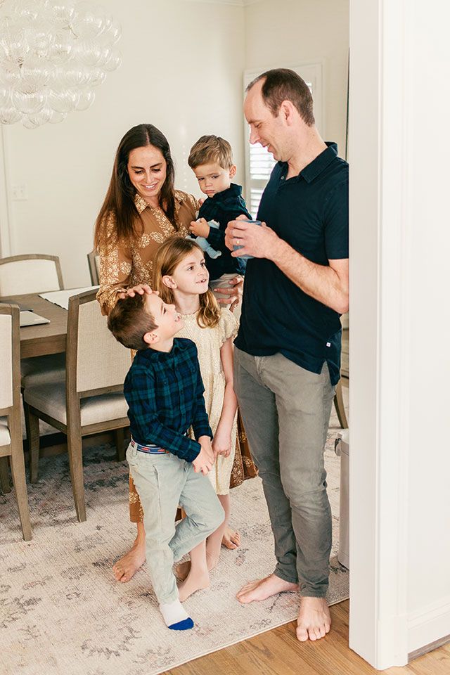A family shares playful moments together in their living room while standing barefoot on a light colored carpet.