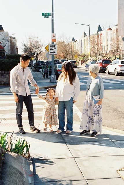 Family of four holding hands at a sunny urban crosswalk, with a young child in a floral dress smiling.