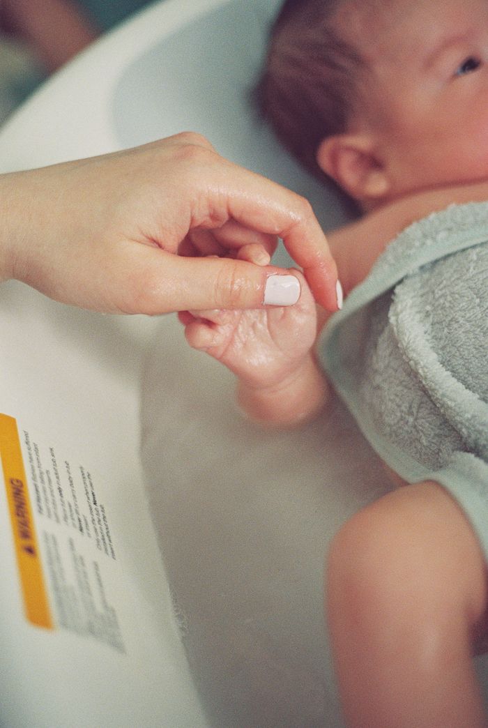 A newborn baby holding an adult's finger during bath time in a white infant tub.