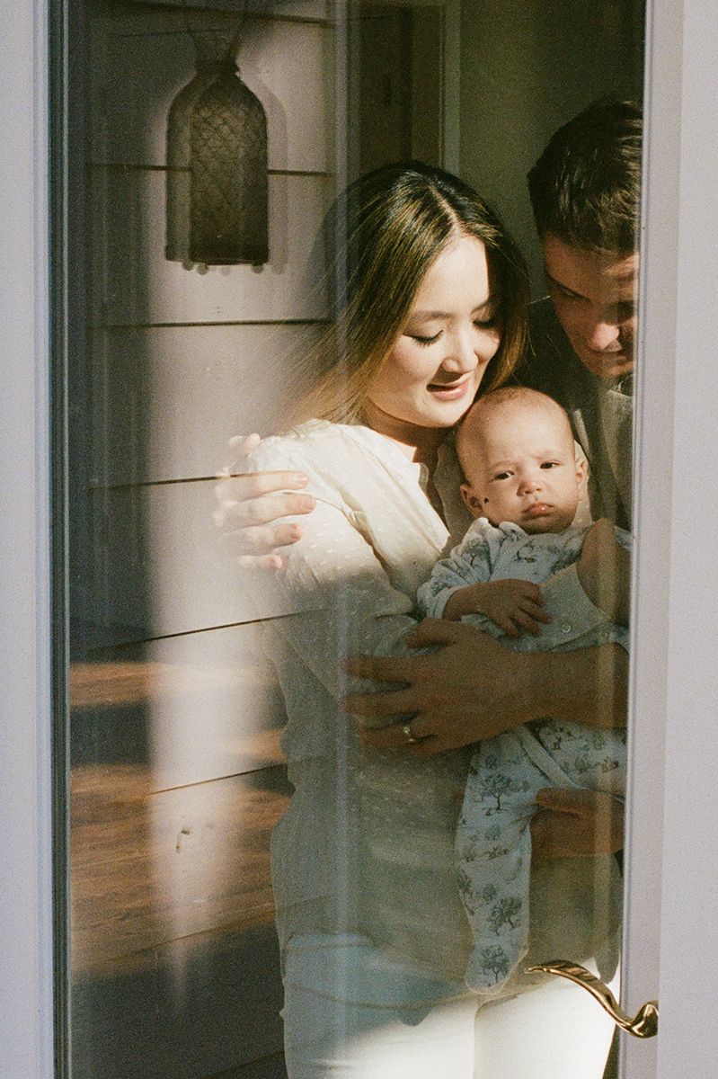 A family stands behind a glass door in soft vintage lighting with their newborn baby.