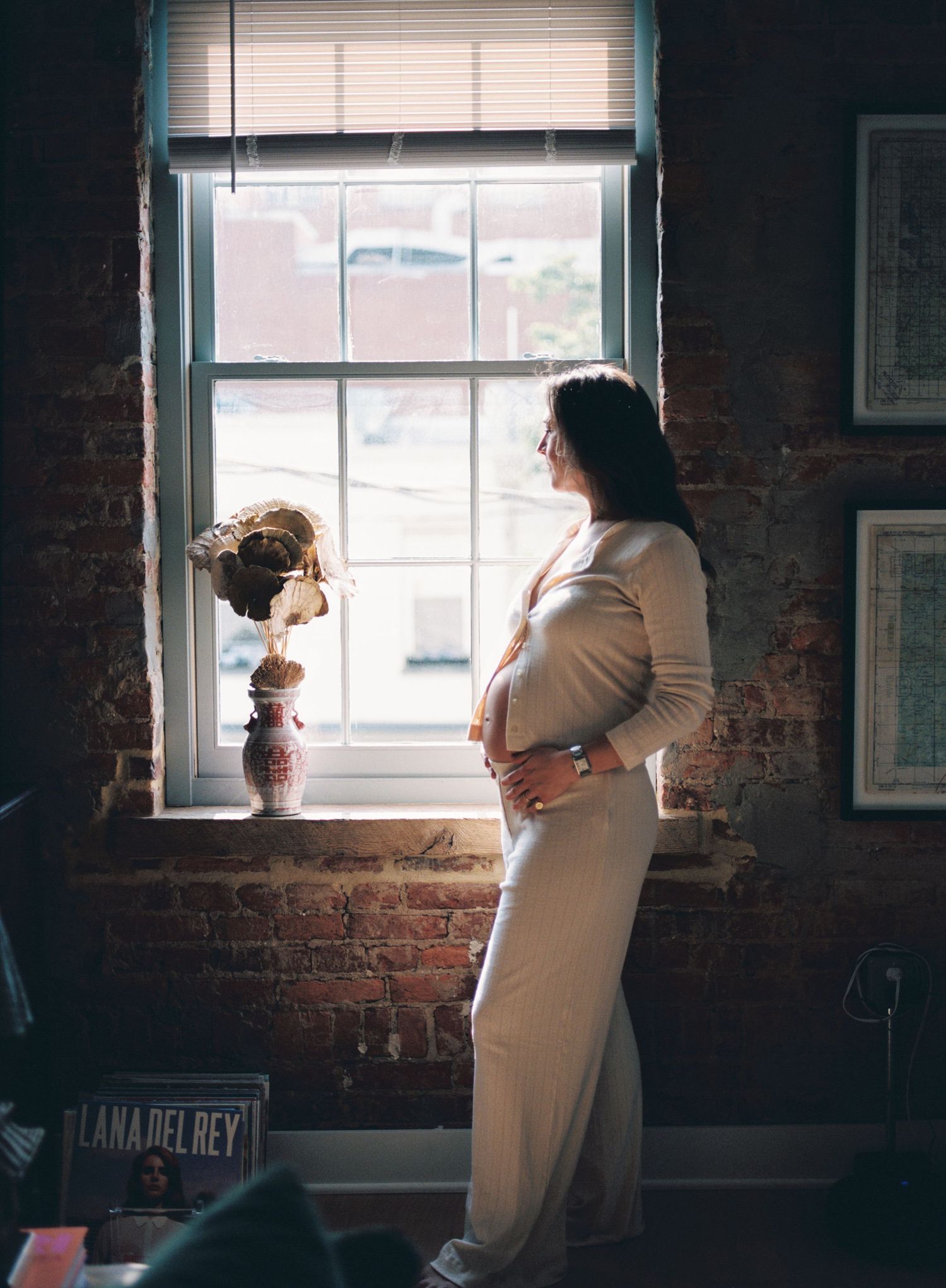 Woman in white dress stands by brick wall window with decorative vase and Lana Del Rey album visible in foreground.